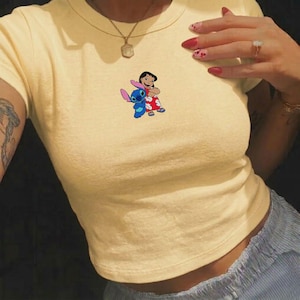 May include: A light yellow crop top featuring a colorful graphic of Lilo and Stitch. The graphic shows Lilo in a red dress and Stitch, a blue alien, standing together. The shirt has short sleeves and a round neckline.