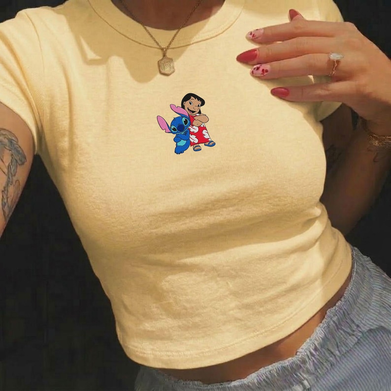 May include: A light yellow crop top featuring a colorful graphic of Lilo and Stitch. The graphic shows Lilo in a red dress and Stitch, a blue alien, standing together. The shirt has short sleeves and a round neckline.