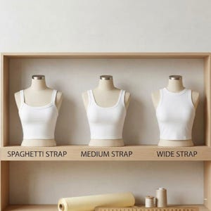 Puede incluir: Tres camisetas sin mangas blancas en maniqu&iacute;es con diferentes estilos de tirantes: tirantes finos, tirantes medianos y tirantes anchos. Las camisetas sin mangas se exhiben en una estanter&iacute;a de madera. El texto debajo indica el estilo de tirantes.