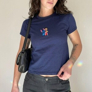 May include: Navy blue t-shirt with a small embroidered design featuring the characters Stitch and Lilo. The shirt has short sleeves and a crew neck. The model is wearing black jeans and a black shoulder bag.