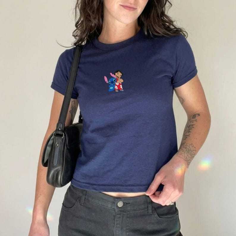 May include: Navy blue t-shirt with a small embroidered design featuring the characters Stitch and Lilo. The shirt has short sleeves and a crew neck. The model is wearing black jeans and a black shoulder bag.