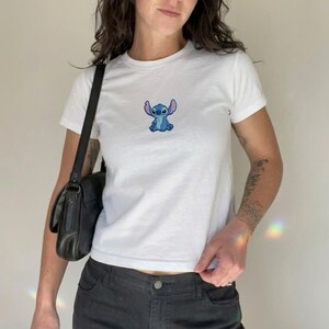 Puede incluir: Camiseta blanca con un peque&ntilde;o bordado azul de Stitch. La camiseta tiene mangas cortas y cuello redondo. El conjunto se completa con pantalones negros y un bolso negro.