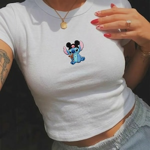 May include: White crop top featuring a cartoon image of a blue alien character with black ears and a red heart. The character is holding a small object. The top has a round neckline and short sleeves.