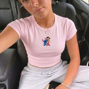 May include: A light pink t-shirt with a crew neck and short sleeves. The shirt features a colorful graphic of the Disney characters Stitch and Lilo. The shirt is worn with gray sweatpants and a gold necklace.