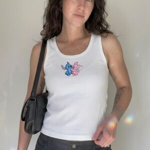 Puede incluir: Camiseta sin mangas blanca acanalada con un gr&aacute;fico de dibujos animados de dos personajes. La camiseta sin mangas tiene cuello redondo y una silueta ajustada. El gr&aacute;fico muestra un personaje azul y rosa abraz&aacute;ndose. Se ve un bolso negro.