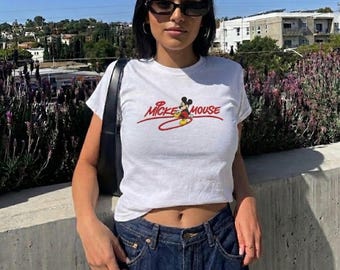Könnte beinhalten: Weißes Crop-Top mit dem Text "Mickey Mouse" in roter Schrift und einer kleinen Mickey Mouse-Grafik. Das Oberteil wird mit dunkelblauen Jeans getragen. Die Person trägt eine schwarze Sonnenbrille und eine schwarze Umhängetasche.