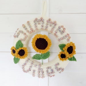 Puede incluir: Una corona de crochet blanca con la palabra "sunshine" escrita en hilo de colores. La corona está decorada con tres girasoles de crochet.