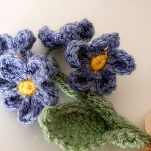 May include: A crocheted bouquet of blue forget-me-not flowers with yellow centers and green leaves.