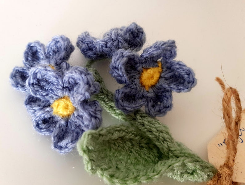May include: A crocheted bouquet of blue forget-me-not flowers with yellow centers and green leaves.