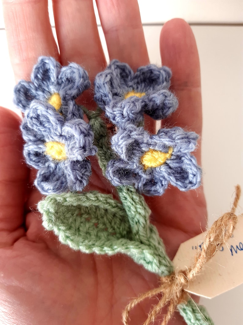 May include: A crocheted bouquet of blue flowers with yellow centers and green leaves. The flowers are tied together with twine and a small tag.