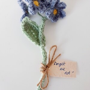 May include: A crocheted forget-me-not flower bouquet with a green stem and blue flowers. The bouquet is tied with twine and has a tag that says "Forget me not".