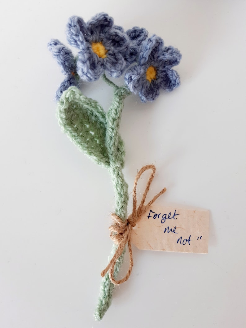 May include: A crocheted forget-me-not flower bouquet with a green stem and blue flowers. The bouquet is tied with twine and has a tag that says "Forget me not".