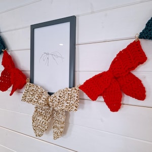 May include: A handmade crochet garland featuring red, cream, and teal bows. The garland is strung with gold-coloured rings and hangs on a white wall. A framed picture with a line drawing is in the centre.