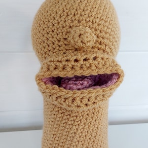 Pu&ograve; includere: Una testa di burattino a uncinetto con un corpo color beige e una bocca rosa. Il burattino ha un piccolo naso rotondo.
