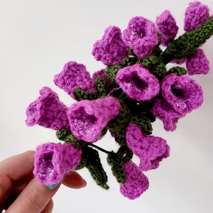 May include: A bouquet of crocheted purple flowers with green leaves. The flowers are shaped like bells and have a textured surface.