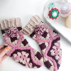 May include: A pair of handmade crochet socks in beige, burgundy, and pink. The socks feature a granny square design with a pom-pom detail and ribbed cuffs. The socks are displayed on a white surface.