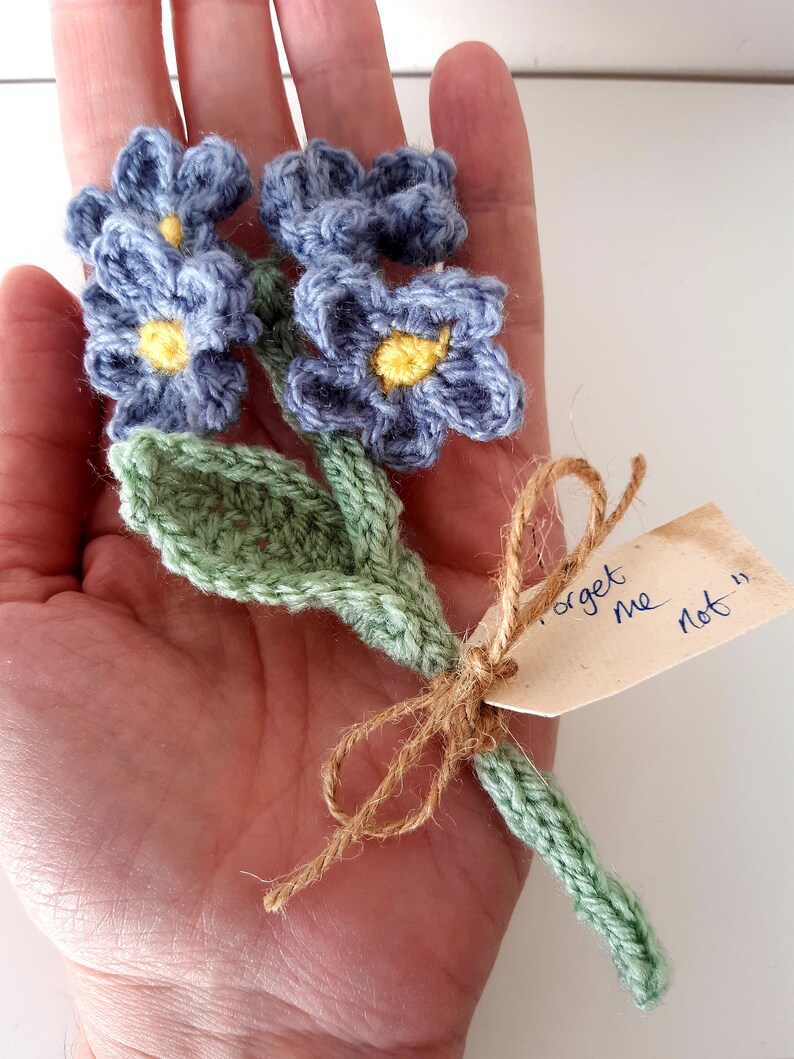 May include: A crocheted bouquet of forget-me-not flowers with green leaves and a tag that says "Forget me not".