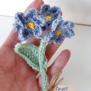 May include: A crocheted bouquet of forget-me-not flowers in shades of blue and green with yellow centers. The flowers are tied together with twine and a small tag that reads "Forget me not".