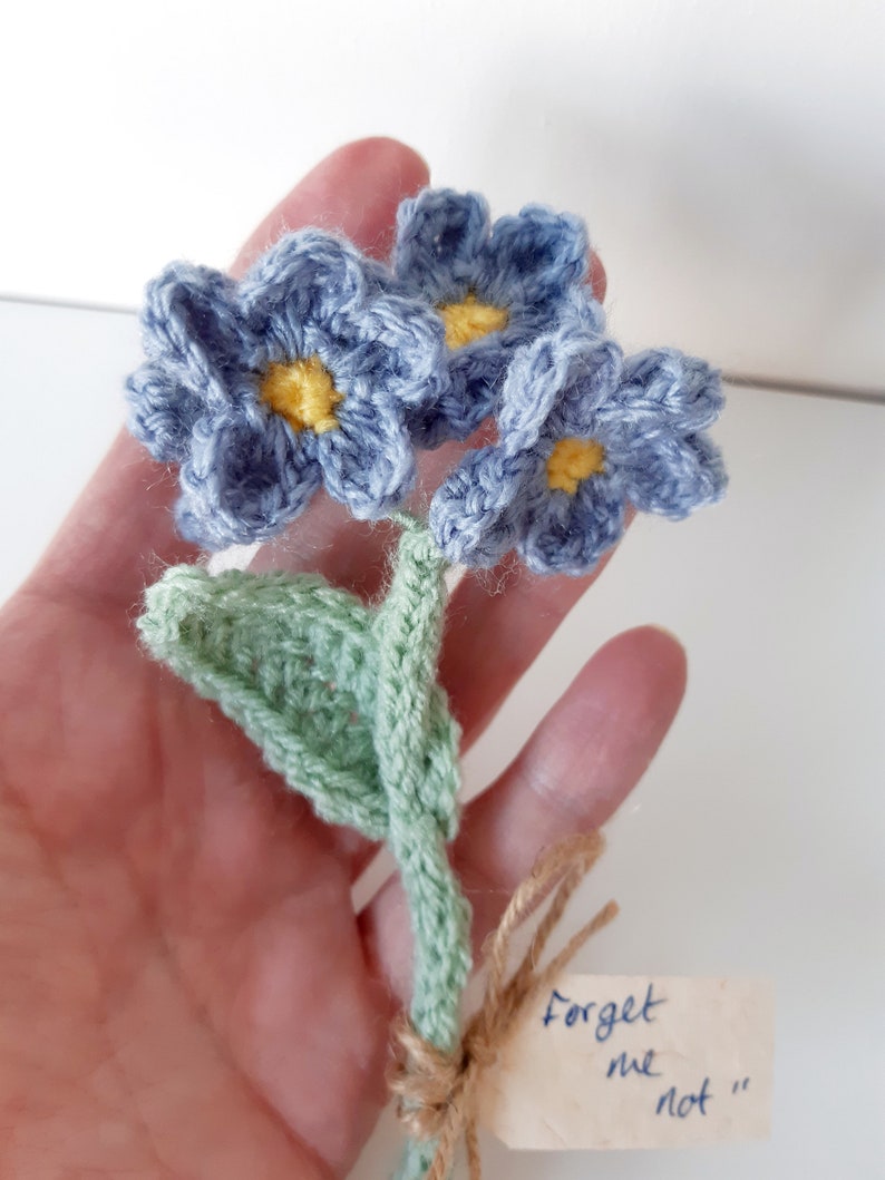 May include: A crocheted bouquet of forget-me-not flowers in shades of blue and green with yellow centers. The flowers are tied together with twine and a small tag that reads "Forget me not".