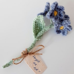 May include: A crocheted forget-me-not flower bouquet with a green stem and blue flowers. The bouquet has a small tag attached with the text "Forget me not".