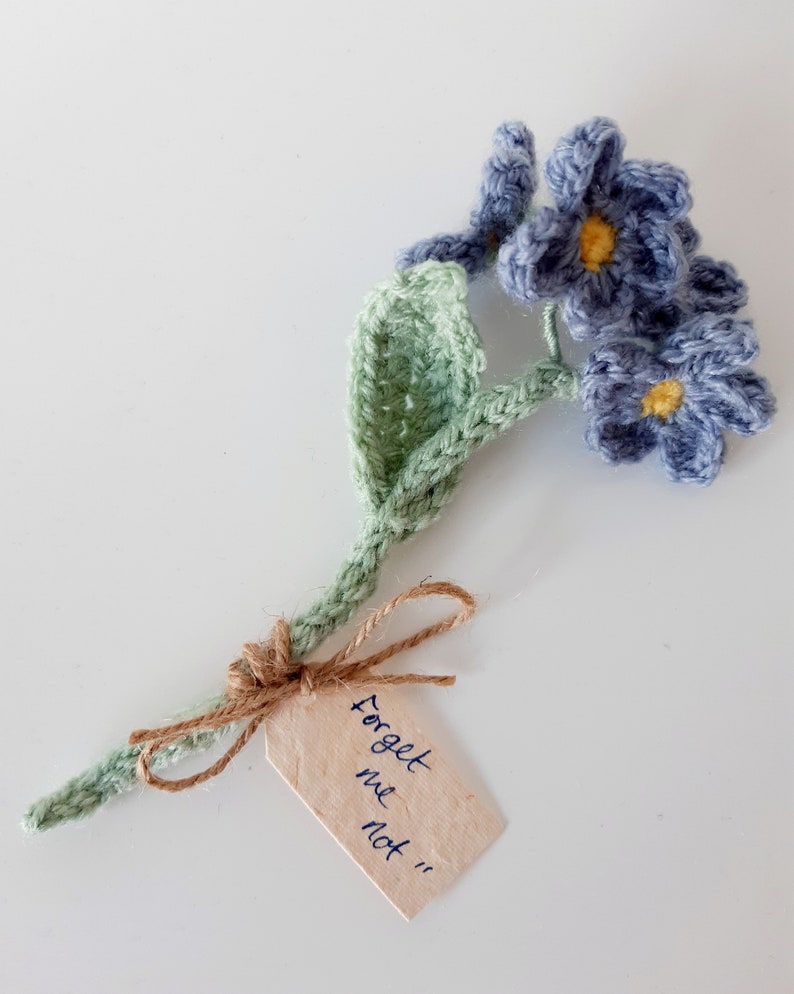 May include: A crocheted forget-me-not flower bouquet with a green stem and blue flowers. The bouquet has a small tag attached with the text "Forget me not".