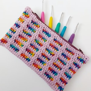 May include: A lavender crochet pouch with a zipper, filled with colorful crochet hooks. The pouch has a textured, geometric pattern with rainbow-colored accents. The crochet hooks have brightly colored handles in pink, yellow, green, blue, and purple.
