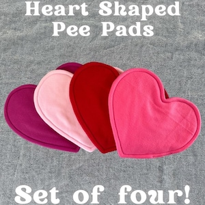 May include: Four heart-shaped pee pads in varying shades of pink and red are displayed against a grey textured backdrop. The text reads "Heart Shaped Pee Pads" and "Set of four!" in white. The Piggy Patch logo is visible.
