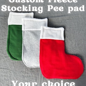 Puede incluir: Tres almohadillas de orina de calcetín de forro polar en verde, gris y rojo, cada una con un puño blanco. El texto dice "Custom Fleece Stocking Pee pad" y "Your choice of colours!". El logotipo de Piggy Patch está en la esquina superior derecha.