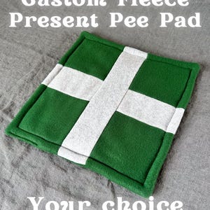 Puede incluir: Una almohadilla de forro polar verde y blanca con un diseño de cruz. La almohadilla tiene un borde verde y se muestra el texto "Custom Fleece Present Pee Pad" y "Your choice of colours!". El logotipo de Piggy Patch está en la esquina superior derecha.