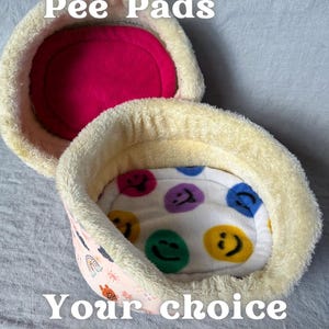 May include: Two Cuddle Cup Pee Pads, one open and one closed. The open pad has a white interior with colorful smiley faces. The closed pad has a bright pink interior. The text reads "Cuddle Cup Pee Pads" and "Your choice of colours!"
