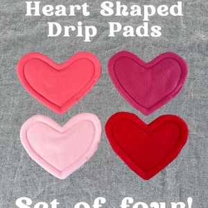 May include: Four heart-shaped drip pads in shades of pink and red are displayed on a grey textured surface. The pads are arranged in a square, with the text "Heart Shaped Drip Pads" and "Set of four!" above and below the pads.