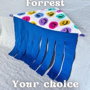 Puede incluir: Un juguete triangular de forro polar con una parte superior blanca con caras sonrientes de colores y una franja azul. Se muestra el texto "Fleece Forrest" y "Your choice of colours!". El logotipo de Piggy Patch está en la esquina superior derecha.