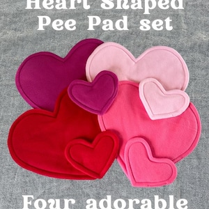 Può includere: Un set di traversine a forma di cuore in varie tonalità di rosa e rosso. L'immagine mostra traversine di diverse dimensioni, con il testo "Heart Shaped Pee Pad set" e "Four adorable colour options!". Il logo The Piggy Patch è in alto a destra.