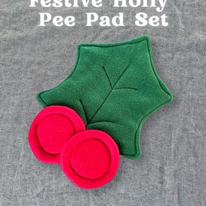 Puede incluir: Un juego de almohadillas de orina de acebo festivo. El juego incluye una hoja de acebo verde y dos almohadillas rojas con forma de bayas. El texto "Festive Holly Pee Pad Set" se muestra en blanco. El logotipo de Piggy Patch está en la esquina superior derecha.