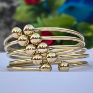 May include: Gold-colored bangle bracelets with a smooth, polished finish. Each bracelet features a series of small, round, gold-colored beads. The bracelets are stacked together, creating a layered effect. The background is blurred, with hints of red, green, and blue.