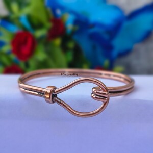 Può includere: Un bracciale rigido in metallo color rame con una chiusura a gancio a forma di goccia al centro. Il bracciale è composto da due fili paralleli che si incontrano in un piccolo connettore cilindrico. Il testo RDIartisanRing è visibile sul bracciale.