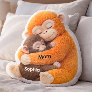 May include: A plush monkey pillow in the shape of a mother monkey hugging a baby monkey. The mother monkey is orange, and the baby monkey is brown. The word "Mom" is printed on the mother monkey, and the name "Sophia" is printed on the baby monkey.