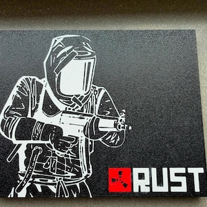 Könnte beinhalten: Eine rechteckige schwarze Platte mit einer weißen grafischen Illustration einer Figur, die einen Schutzhelm trägt und eine Schusswaffe hält. Das Wort RUST ist in weißen Großbuchstaben neben einem roten Quadrat aufgedruckt.