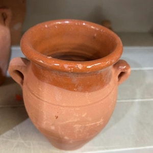 Peut inclure: Un petit vase de couleur terre cuite avec deux petites anses. Le bord et les anses sont d'une nuance de brun plus foncée. Le vase a un corps arrondi et une large ouverture. Le vase est posé sur une étagère.