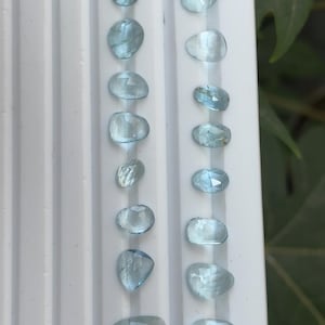 Peut inclure: Une collection de pierres précieuses bleu clair de formes et tailles variées, disposées en deux rangées verticales sur une surface blanche. Les pierres ont une qualité translucide, avec des facettes qui captent la lumière. L'arrière-plan est flou.