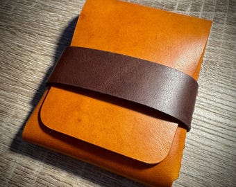 May include: A rectangular leather wallet featuring a smooth tan main body and a contrasting dark brown leather strap wrapped horizontally across the center. The wallet has a clean, folded construction with visible grain and stitched edges.