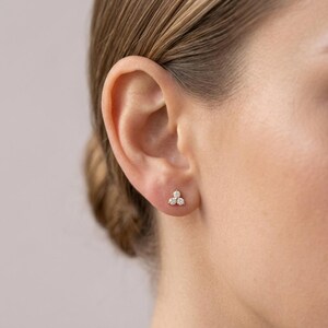 May include: A gold stud earring featuring three small, round, clear stones arranged in a triangular cluster. The earring is positioned in the lower lobe of an ear, showcasing a polished metal setting that holds the stones securely in place.