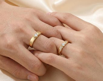 May include: Two gold rings featuring a central round-cut clear stone. One ring has a smooth, polished band, while the other features a channel-set band with small clear stones along the sides, both showcasing a bright, reflective metallic finish.