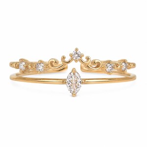 Puede incluir: Un anillo abierto de tono dorado con una piedra central transparente de corte marquesa en la banda inferior y una banda superior decorativa con volutas. La banda superior está adornada con cinco pequeñas piedras redondas transparentes.