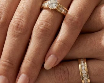 May include: A gold engagement ring featuring a large round brilliant-cut center diamond with smaller diamonds set along the band, paired with a matching gold wedding band featuring a channel of round diamonds set across the center of the band.