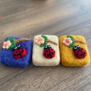 Könnte beinhalten: Drei gefilzte Seifenstücke in Blau, Creme und Gelb, jeweils mit einem roten Marienkäfer und rosa Blüten auf einem Zweig verziert. Die Marienkäfer haben schwarze Punkte und weiße Augen. Die Seifen sind rechteckig und liegen auf einer Holzoberfläche.
