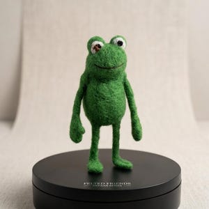 Puede incluir: Figura de rana verde hecha a mano con fieltro, con una expresión sonriente. La rana tiene ojos negros y blancos y está sobre una base circular negra. La base tiene el texto "FELTED FRIENDS ARTISAN SERIES".