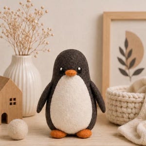 Op de afbeelding: Een handgemaakte pinguïn knuffel met een donkergrijze body, witte buik en oranje snavel en voeten. De pinguïn staat op een houten oppervlak. De achtergrond bevat een witte vaas met gedroogde bloemen, een klein houten huisje en een ingelijste print.