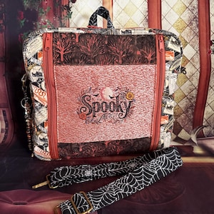 May include: A rectangular fabric bag featuring a central panel with the embroidered word Spooky, surrounded by floral and bat motifs. The bag has a dark tree pattern, orange zippers, and a detachable black strap with a white spiderweb print.