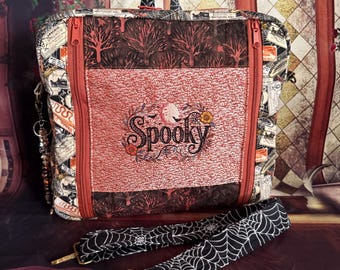 May include: A rectangular fabric bag featuring a central panel with the embroidered word Spooky, surrounded by floral and bat motifs. The bag has a dark tree pattern, orange zips, and a detachable black strap with a white spiderweb print.